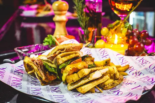 Club Sandwich & Fresh French Fries on a Pose branded paper on a plate; a lit candle, a pepper grinder, grapes and a glass of white wine in the background