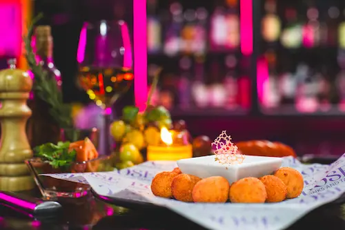 Serrano Ham & Bechamel Croquettes served with Secret dip on a Pose branded paper on a plate; a lit candle, a pepper grinder, grapes and a glass of white wine in the background