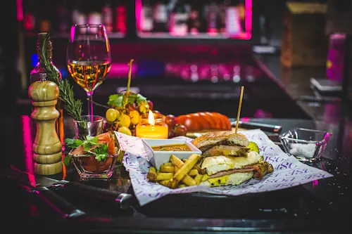 Cuban Sandwich, dip & freshly cooked French Fries on a Pose branded paper on a plate; a lit candle, a pepper grinder, grapes and a glass of white wine in the background