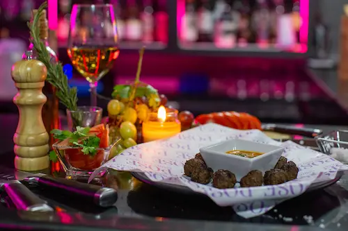 Meatballs & Secret dip on a Pose branded paper on a plate; cutlery, a lit candle, a pepper grinder, grapes and a glass of white wine in the background