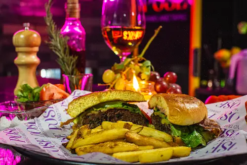 Beef & Cheese Burger & freshly cooked French Fries on a Pose branded paper on a plate; a lit candle, a pepper grinder, grapes and a glass of white wine in the background